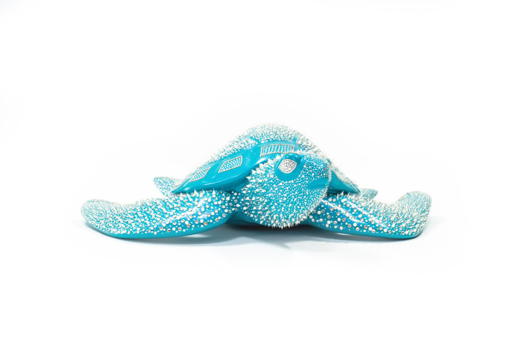 Turtle Turquoise – Eddy Maniez De Turtle Turquoise van Eddy Maniez is een meesterlijke sculptuur die onmiddellijk de aandacht grijpt. Met zijn levendige turquoise kleur en duizenden silicone dots gecombineerd met Swarovski kristallen, ademt dit werk kracht én verfijning. Het fonkelt in het licht en verandert voortdurend van uitstraling, waardoor elke blik een nieuwe ervaring biedt. Deze zeeschildpad symboliseert vrijheid, bescherming en tijdloosheid. Het werk laat zien hoe natuur en luxe elkaar kunnen versterken, zonder hun essentie te verliezen. Door de stralende turquoise tint roept Turtle Turquoise gevoelens van rust en harmonie op, terwijl de dynamische textuur juist energie en beweging uitstraalt. Dit contrast maakt de sculptuur zowel een krachtig middelpunt als een verfijnd kunstwerk dat een ruimte transformeert. Het is een kunstwerk dat niet enkel bekeken wordt, maar ook emoties losmaakt en een blijvende indruk nalaat.