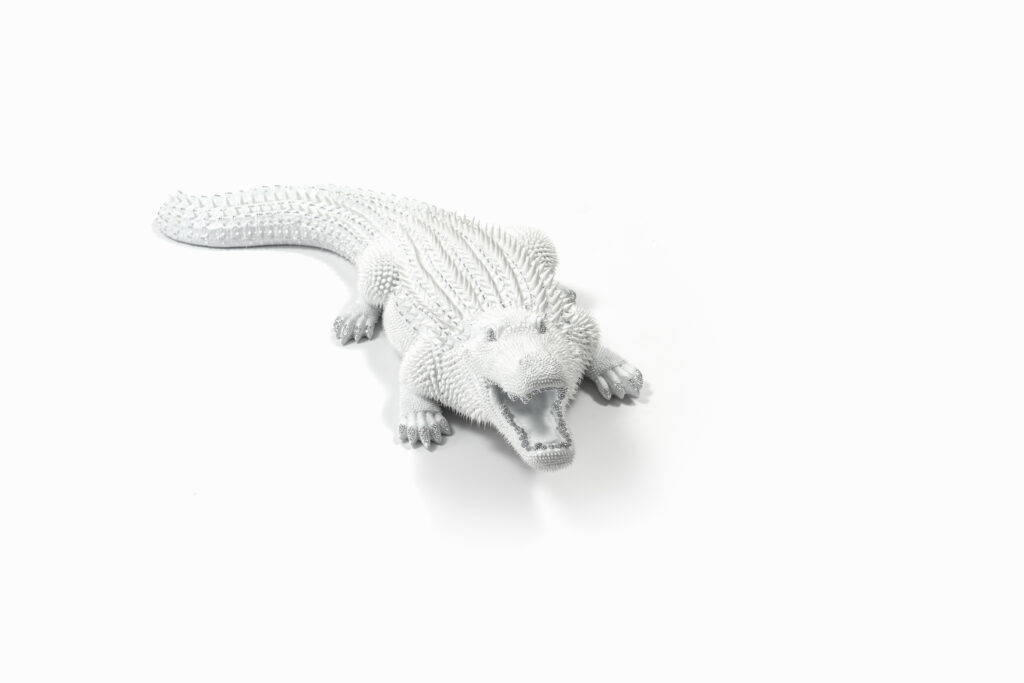 Crocodile White – Eddy Maniez Crocodile White van Eddy Maniez is een indrukwekkend kunstwerk dat kracht, luxe en verfijning belichaamt. De sculptuur toont een krokodil in volle beweging, zijn bek wijd geopend alsof hij midden in een krachtige uitbarsting is vastgelegd. Het volledige oppervlak is bedekt met silicone dots en fonkelende Swarovski kristallen, waardoor de krokodil een unieke, bijna futuristische uitstraling krijgt. De witte kleur versterkt het gevoel van zuiverheid en elegantie, terwijl de glinsteringen zorgen voor een aura van extravagantie. Bovendien benadrukt Crocodile White de symboliek van de krokodil: oeroude kracht, bescherming en een ontembare levensenergie. Door dit dier in wit uit te voeren, doorbreekt Maniez het traditionele beeld van de krokodil als een donker, gevaarlijk roofdier. Hij transformeert het tot een luxueus icoon dat zowel gevaar als schoonheid in zich draagt. Dit contrast maakt het werk spannend, intrigerend en onvergetelijk. De details zijn adembenemend. Van de scherpe tanden tot de textuur van de huid, elk element is zorgvuldig bewerkt en voorzien van kristallen accenten. Het resultaat is een sculptuur die zowel artistiek als technisch verbluffend is. Crocodile White is meer dan een object: het is een statement piece dat iedere ruimte domineert en onmiddellijk de aandacht trekt.