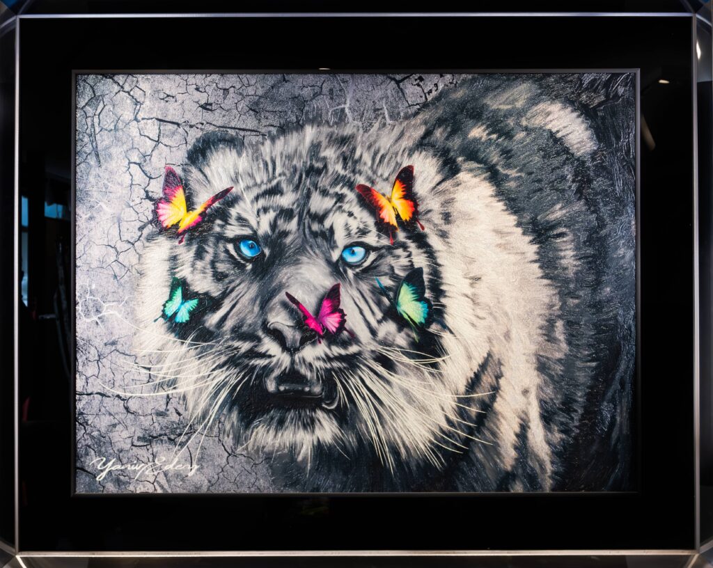 Yaniv Edery – Big Lion Multicolor Dit indrukwekkende kunstwerk van Yaniv Edery toont een majestueuze grote kat die de kijker recht in de ogen kijkt. De doordringende blauwe ogen trekken onmiddellijk de aandacht en geven het werk een intens en mysterieus karakter. Rond het gezicht fladderen kleurrijke vlinders in rood, groen en oranje. Zij zorgen voor een verrassend contrast en brengen luchtigheid en hoop in het krachtige beeld. De achtergrond, uitgevoerd in een ruwe grijstint met scheuren en textuur, versterkt de spanning. Het grimmige decor benadrukt de kracht en de kwetsbaarheid van het dier. De felle kleuren van de vlinders lijken te dansen op de donkere ondergrond en creëren een harmonieus samenspel van kracht en zachtheid. Dit maakt het kunstwerk zowel dreigend als poëtisch. Edery laat met dit werk zien hoe tegenstellingen elkaar versterken. De grote kat staat symbool voor macht, instinct en natuur, terwijl de vlinders lichtheid, transformatie en vrijheid uitbeelden. Deze balans tussen kracht en tederheid maakt het kunstwerk uniek en gelaagd. Het vertelt een verhaal over veerkracht en schoonheid te midden van een harde wereld. Elk detail, van de glans in de ogen tot de delicate vleugels van de vlinders, is zorgvuldig uitgewerkt. Big Lion Multicolor is meer dan een visueel spektakel: het is een symbool van hoop, moed en innerlijke kracht. Het kunstwerk weet emoties los te maken en nodigt uit tot reflectie.