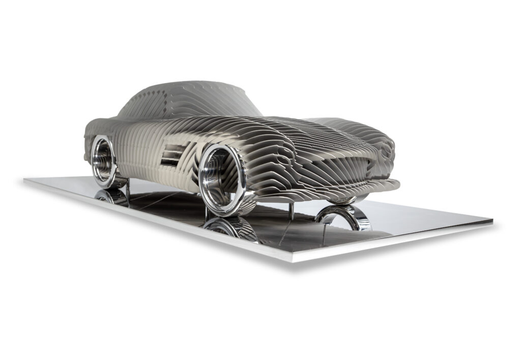 Antoine Dufilho – Mercedes 300 SL Streamline Crème Het kunstwerk Mercedes 300 SL Streamline Crème van Antoine Dufilho is een ode aan snelheid en elegantie. Het werk toont de iconische Mercedes 300 SL in een futuristische interpretatie. De sculptuur bestaat uit gelaagde, gestroomlijnde vormen die ritme en beweging suggereren. Elke lijn benadrukt de vloeiende dynamiek van de auto. Het samenspel van open ruimte en volume geeft het werk een lichtheid, alsof het zweeft. De sculptuur trekt direct de aandacht. Strakke lijnen wisselen af met subtiele rondingen, waardoor een harmonieus beeld ontstaat. Het gebruik van crème- en metaaltinten versterkt het gevoel van luxe en raffinement. Bovendien wordt de auto getoond als meer dan een voertuig: het is een symbool van vooruitgang, design en techniek. Dufilho weet met dit werk een balans te vinden tussen abstractie en herkenning. Van dichtbij zie je de ritmische structuren. Van verder weg herken je duidelijk de iconische Mercedes. Daardoor krijgt het werk meerdere lagen en nodigt het uit tot langer kijken. Het is zowel modern als tijdloos.