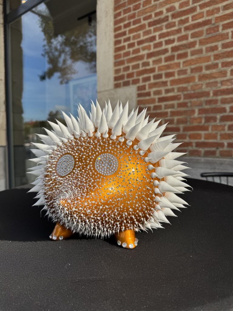 Hedgehog Golden Yellow – Eddy Maniez Hedgehog Golden Yellow van Eddy Maniez is een speels en extravagant kunstwerk waarin natuur en luxe samenkomen. Deze sculptuur toont een egeltje dat volledig is omgevormd tot een oogverblindend designobject. Het lichaam is uitgevoerd in een glanzende goudgele tint, terwijl talloze kristallen het oppervlak laten fonkelen bij elke lichtinval. De scherpe witte stekels contrasteren krachtig met de warme kleur van de basis, wat het werk een dynamische en energieke uitstraling geeft. Bovendien straalt Hedgehog Golden Yellow zowel kracht als tederheid uit. Het egeltje, normaal symbool voor kwetsbaarheid en bescherming, wordt door Maniez verheven tot een glamoureus icoon. De combinatie van zachte rondingen, glinsterende details en scherpe stekels maakt het beeld spannend en intrigerend. Het werk is niet alleen een eerbetoon aan de natuur, maar ook een expressie van luxe en verbeeldingskracht. Het kunstwerk nodigt uit tot bewondering en nieuwsgierigheid. De sprankeling van de kristallen en de speelse vorm zorgen ervoor dat het stuk direct de aandacht trekt en een ruimte transformeert. Het is meer dan een decoratief object: het is een krachtig statement van stijl, lef en extravagantie.