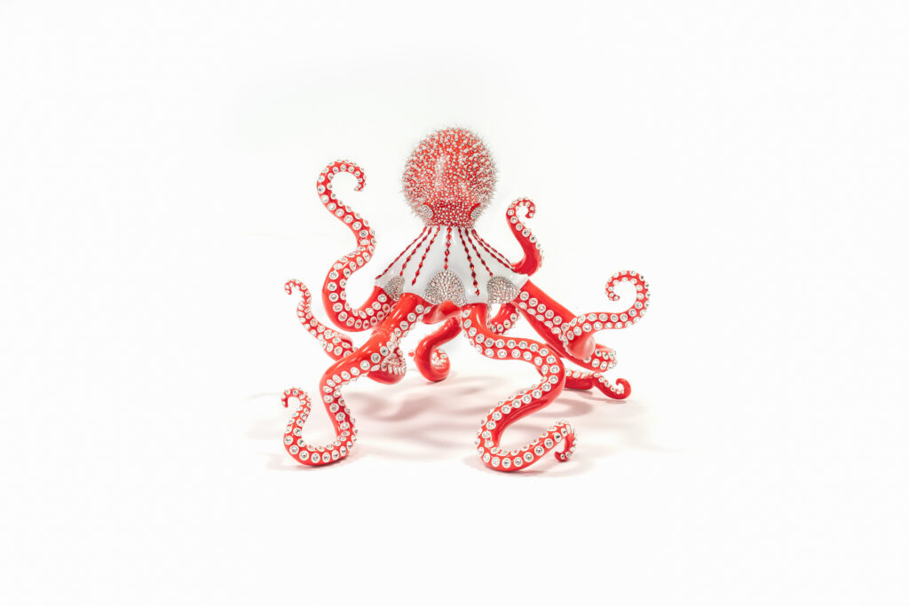 Poulpe Red and White – Eddy Maniez Poulpe Red and White van Eddy Maniez is een meesterlijk kunstwerk waarin kracht, luxe en speelsheid samenkomen. De sculptuur toont een imposante octopus, uitgevoerd in een opvallende combinatie van felrood en helder wit. De tentakels kronkelen sierlijk in verschillende richtingen, wat de sculptuur een dynamische en bijna dansende uitstraling geeft. Elk oppervlak is rijkelijk bezet met Swarovski kristallen en silicone dots, die het kunstwerk laten fonkelen bij iedere lichtinval. Bovendien benadrukt Poulpe Red and White de symboliek van de octopus: mysterie, intelligentie en aanpassingsvermogen. Door de combinatie van kleuren en materialen geeft Maniez dit zeedier een glamoureuze, moderne interpretatie. Het resultaat is een sculptuur die niet alleen de kracht van de natuur viert, maar ook een luxueus statement vormt. Het contrast tussen rood en wit versterkt de visuele impact, terwijl de kristallen een gevoel van elegantie en overdaad toevoegen. Dit werk overstijgt traditionele sculptuur. Het is een iconisch object dat iedere ruimte onmiddellijk transformeert. Poulpe Red and White nodigt uit tot bewondering en roept tegelijkertijd een gevoel van energie en verfijning op. Het is meer dan een kunstwerk; het is een blikvanger, een gespreksonderwerp en een symbool van extravagantie.
