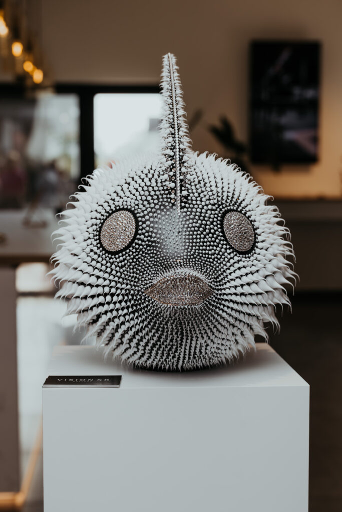Moonfish Black – Eddy Maniez Moonfish Black van Eddy Maniez is een adembenemende sculptuur waarin natuur, luxe en verbeeldingskracht samenkomen. Het werk toont een kogelvis, vormgegeven met een ronde, bijna buitenaardse uitstraling. Het lichaam is diepzwart en bezet met talloze siliconen dots en Swarovski kristallen, die ritmisch over het oppervlak verspreid zijn. Dit patroon geeft de sculptuur niet alleen textuur, maar ook een hypnotiserende dynamiek. De ogen en mond zijn rijkelijk ingelegd met fonkelende Swarovski kristallen. Hierdoor ontstaat een intrigerende spanning tussen speelsheid en extravagantie. Bovendien benadrukt dit contrast de mysterieuze schoonheid van de maanvis, een dier dat symbool staat voor zeldzaamheid en elegantie. Door zijn bijzondere vorm en luxueuze afwerking transformeert Moonfish Black in een echt statement piece dat onmiddellijk de aandacht trekt. Dit kunstwerk is meer dan alleen een representatie van een zeedier. Het is een iconisch object waarin de kwetsbaarheid van de natuur en de overdaad van luxe hand in hand gaan. De ronde vorm, glanzende details en ritmische verspreiding van dots en kristallen maken het beeld tegelijk speels en majestueus. Het nodigt de toeschouwer uit om dichterbij te komen, de details te ontdekken en zich te verliezen in het hypnotische patroon van licht en reflectie. Moonfish Black is een ultieme weerspiegeling van Maniez’ talent om het gewone te verheffen tot iets buitengewoons.
