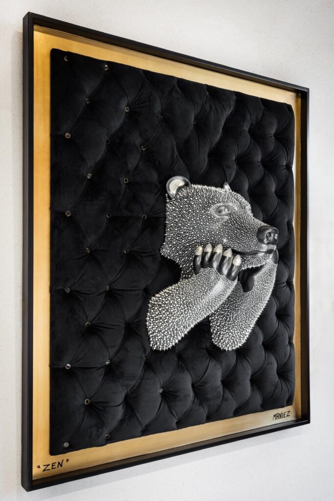 Tableau ZEN Black – Eddy Maniez Tableau ZEN Black van Eddy Maniez is een indrukwekkend kunstwerk dat rust en kracht in één beeld weet te verenigen. Centraal staat de kop en voorpoten van een beer, uitgevoerd in zwart en rijkelijk versierd met fonkelende kristallen. Het dier leunt ontspannen met zijn kop op zijn poten, alsof hij volledig in een staat van meditatie verkeert. Dit straalt kalmte uit, maar behoudt tegelijkertijd een krachtige aanwezigheid. De beer is geplaatst tegen een luxueuze, zwart gewatteerde achtergrond, omlijst met een gouden rand. Deze keuze voor materialen en kleuren versterkt de majestueuze uitstraling van het werk. Het contrast tussen het zachte, matte fluweel en de sprankelende kristallen zorgt voor een fascinerende spanning. Bovendien benadrukt dit contrast het concept van balans: kracht en zachtheid, luxe en natuur, stilte en energie. Tableau ZEN Black is meer dan een kunstwerk; het is een uitnodiging om stil te staan, te ademen en sereniteit te ervaren in een wereld vol prikkels. Door de combinatie van een iconisch dier met een extravagante, moderne afwerking creëert Maniez een werk dat zowel harmonieus als adembenemend is.
