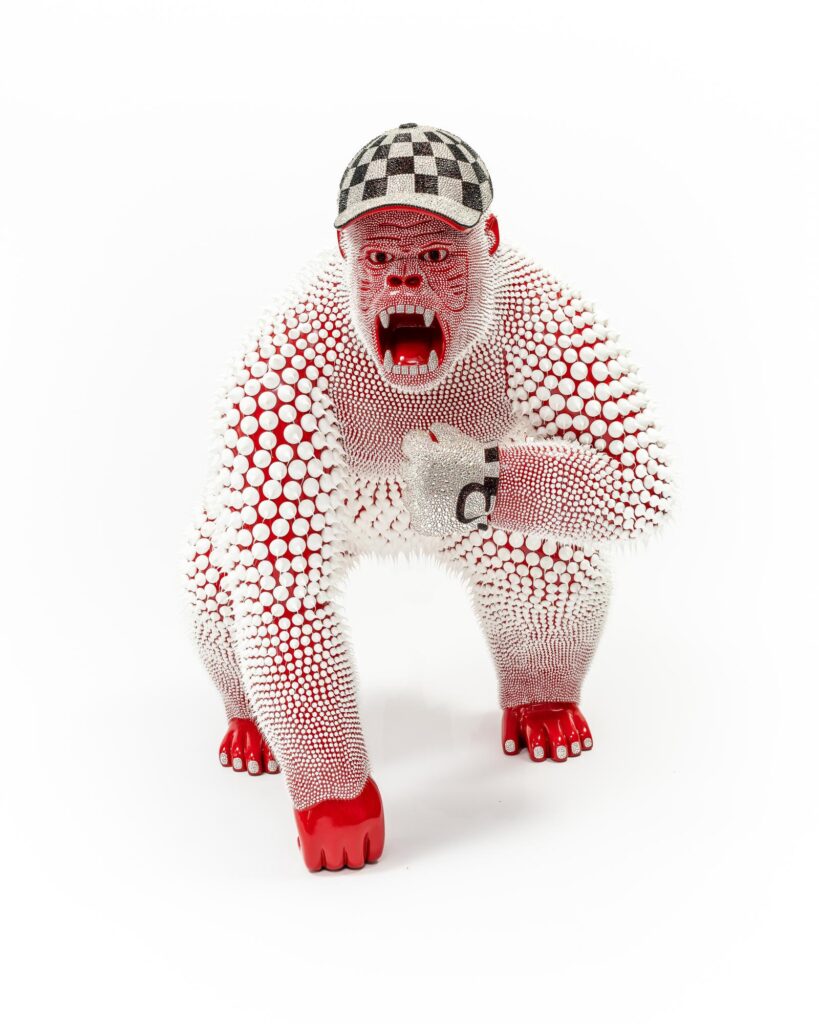 Eddy Maniez – Gorilla Cap and Gloves Red De Gorilla Cap and Gloves Red van Eddy Maniez is een krachtig kunstwerk dat onmiddellijk een onuitwisbare indruk maakt. Deze sculptuur combineert rauwe energie met extravagante verfijning. De gorilla, gehuld in een explosieve rood-witte huid van honderden silicone dots en glinsterende Swarovski kristallen, straalt pure vitaliteit uit. Met zijn agressieve houding en open mond lijkt hij klaar om de toeschouwer te confronteren. Tegelijk voegen de gedetailleerde handschoenen en de zwart-witte geruite pet een speelse, moderne twist toe. Dit contrast maakt het werk niet alleen intrigerend, maar ook prikkelend en vernieuwend. De texturen, het kleurgebruik en de luxueuze kristallen zorgen voor een hypnotiserende gelaagdheid. Het beeld is niet zomaar decoratief; het is een manifest van kracht, humor en hedendaagse kunst in één. Met zijn imposante uitstraling en extravagante details overstijgt de Gorilla Cap and Gloves Red de grens tussen klassieke beeldhouwkunst en moderne popart.