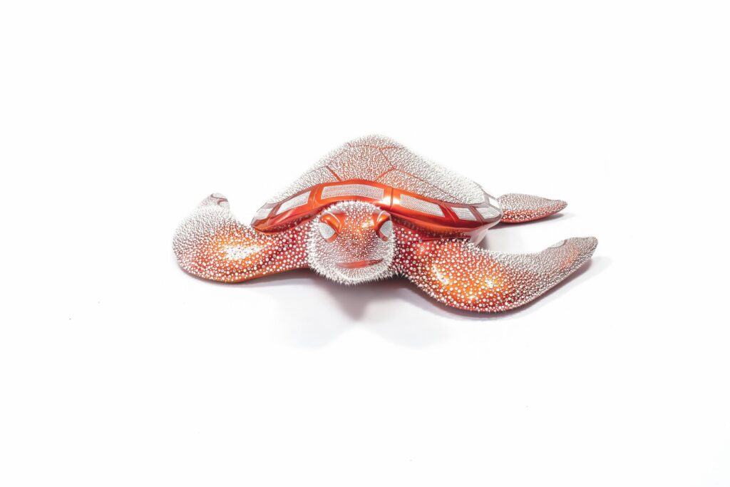Turtle Orange Candy – Eddy Maniez Turtle Orange Candy van Eddy Maniez is een oogverblindende sculptuur die de kracht van de natuur en de pracht van luxe samenbrengt. De zeeschildpad, symbool van wijsheid, geduld en bescherming, is uitgevoerd in een levendige oranje kleur die doet denken aan glanzend snoepgoed. Het oppervlak is rijkelijk bezet met silicone dots en Swarovski kristallen, die sprankelen bij iedere beweging van het licht. Deze combinatie maakt het kunstwerk speels en tegelijkertijd majestueus. Bovendien benadrukt Turtle Orange Candy de dualiteit tussen zachtheid en kracht. De schildpad, vaak gezien als een kalm en sereen wezen, krijgt in dit werk een extravagante en moderne uitstraling. De lange vinnen en het sierlijk gevormde schild worden geaccentueerd door de glanzende kristallen, waardoor het dier bijna mythisch aanvoelt. Het kunstwerk overstijgt daarmee een realistische weergave en verandert in een luxueus icoon dat schoonheid, harmonie en overdaad uitstraalt. Het gebruik van oranje als hoofdkleur staat symbool voor energie, vitaliteit en optimisme. Samen met de kristallen vormt het een hypnotiserend geheel dat de aandacht onmiddellijk naar zich toe trekt. Turtle Orange Candy is niet slechts een sculptuur, maar een krachtig statement piece dat iedere ruimte transformeert in een plek van luxe en verbeelding.