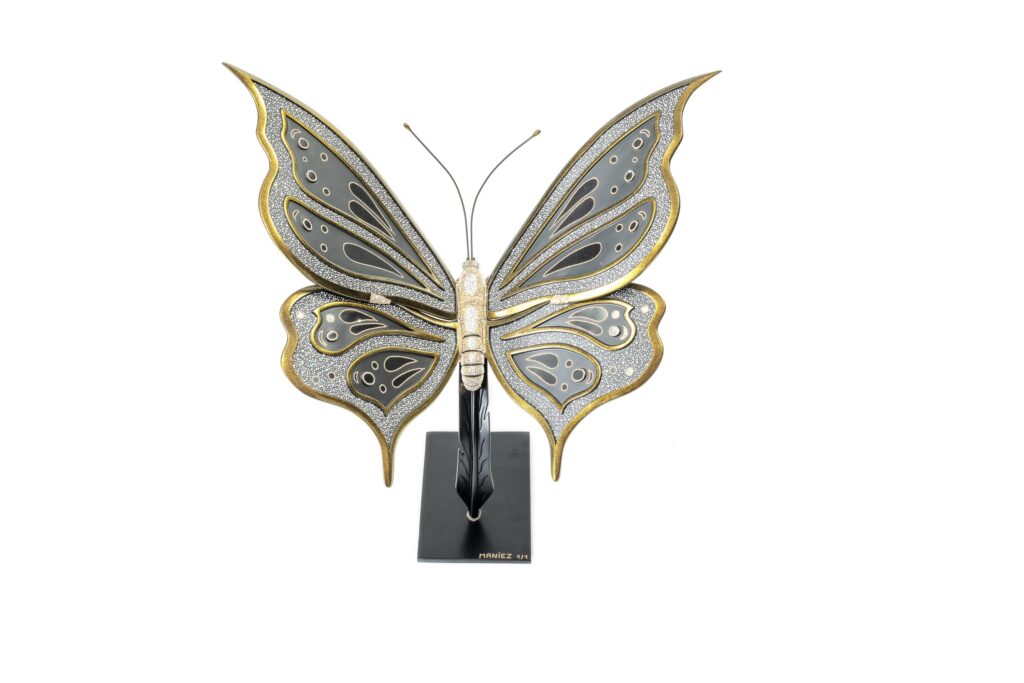 Butterfly Black and Gold – Eddy Maniez Butterfly Black and Gold van Eddy Maniez is een adembenemend kunstwerk waarin elegantie, luxe en symboliek samenkomen. Deze sculptuur toont een sierlijke vlinder, uitgevoerd in diepe zwarte tinten en rijk afgewerkt met gouden accenten. De vleugels zijn ingelegd met talloze fonkelende Swarovski kristallen, waardoor het werk schittert zodra het licht erop valt. Het contrast tussen het donkere zwart en het warme goud benadrukt de gracieuze vormen en geeft de vlinder een bijna koninklijke uitstraling. Bovendien is de vlinder al eeuwenlang een krachtig symbool voor transformatie, vrijheid en schoonheid. In Butterfly Black and Gold verheft Maniez dit symbool tot een luxueus icoon. De perfecte balans tussen verfijnde details en sprankelende weelde maakt het kunstwerk zowel modern als tijdloos. De sculptuur is niet alleen een ode aan de natuur, maar ook een expressie van elegantie en kracht. Dit kunstwerk trekt onmiddellijk de aandacht. De ranke vormen, rijke materialen en schitterende details zorgen ervoor dat het een echte eyecatcher is in iedere ruimte. Butterfly Black and Gold is meer dan decoratie; het is een statement piece dat verfijning en luxe belichaamt.